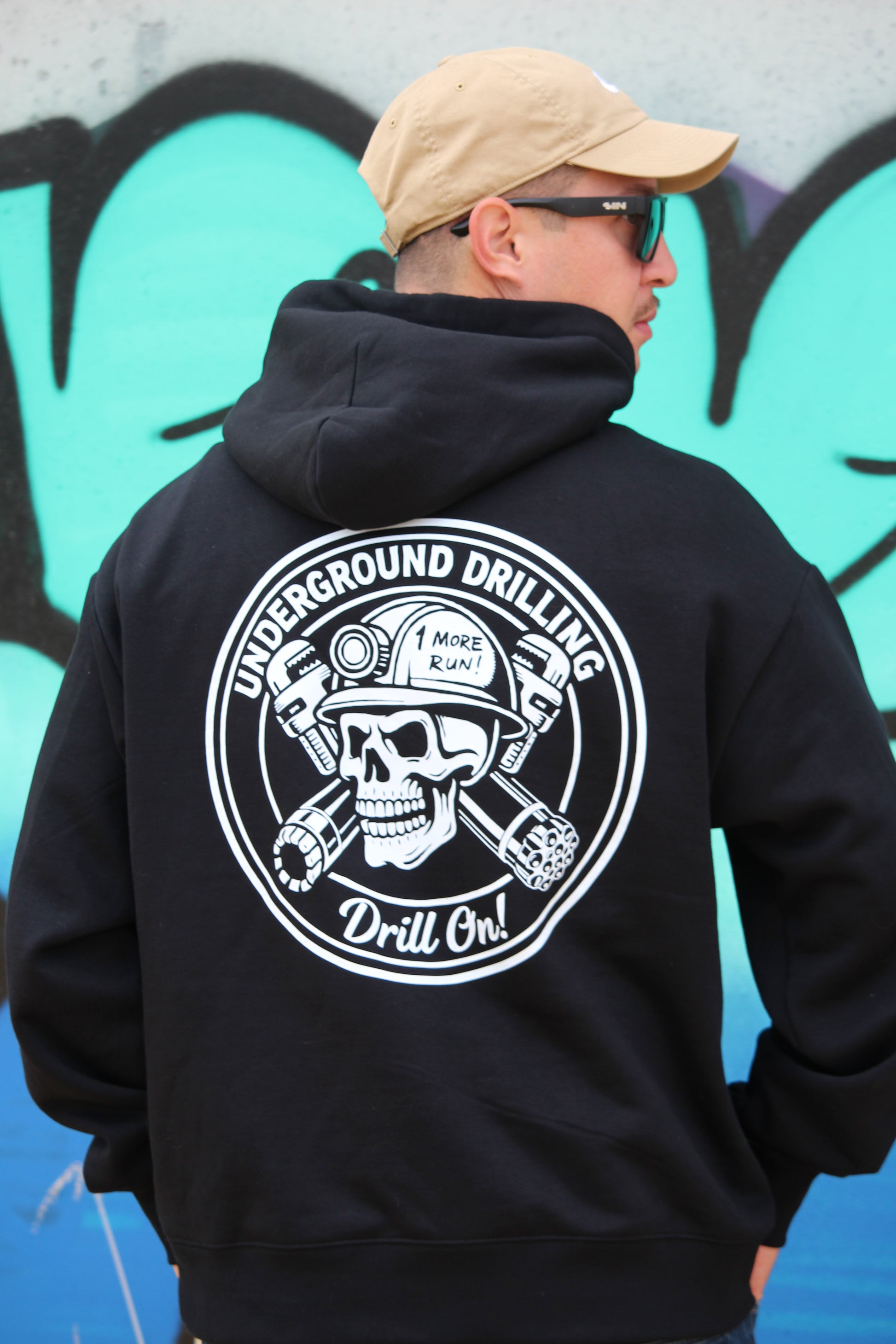 Person wearing a black hoodie with a white logo against a graffiti-covered wall.