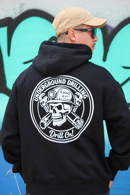 Person wearing a black hoodie with a white logo against a graffiti-covered wall.